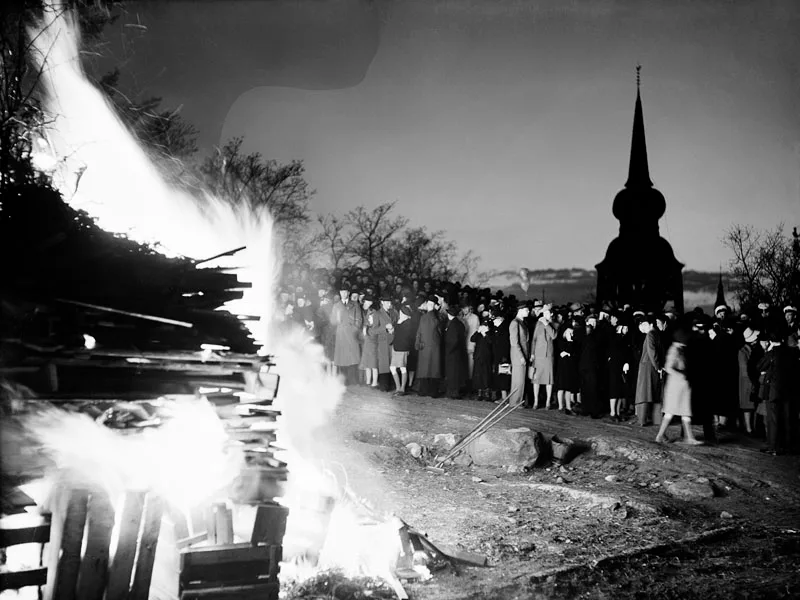Fogueira alta à esquerda e uma multidão de sobretudo no terreno do Skansen, com a torre de uma igreja de madeira ao fundo, na noite de Valborg de 1939.