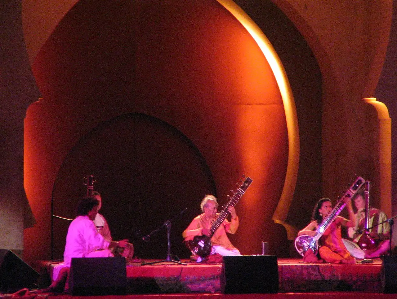 Músicos tocando sitar num palco iluminado em tons de vermelho, sob um grande arco de estilo mourisco, no Festival de Música Sacra de Fez.