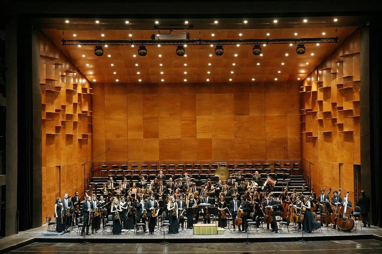Orchestre au complet sur une scène en bois clair sous un plafond de projecteurs, lors d’un concert du Maggio Musicale Fiorentino.