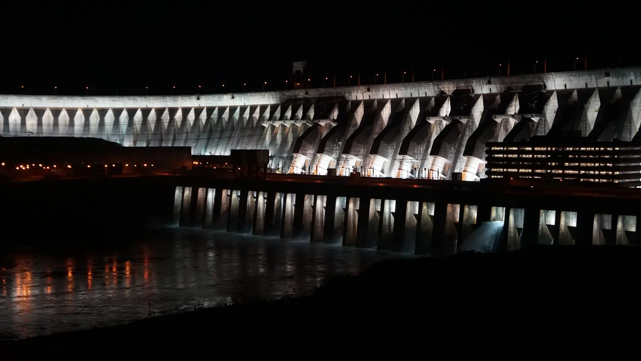 A barragem e o vertedouro de Itaipu iluminados de branco à noite, em 2 de novembro de 2012, com uma comporta aberta e o reflexo na água.