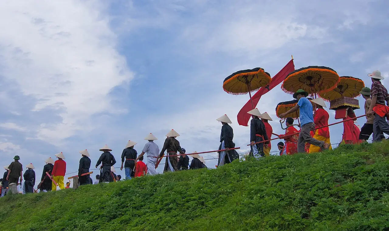 Fila de participantes com chapéus cônicos e sombrinhas amarelas e vermelhas caminhando ao longo de um aterro gramado durante o festival de Gióng.