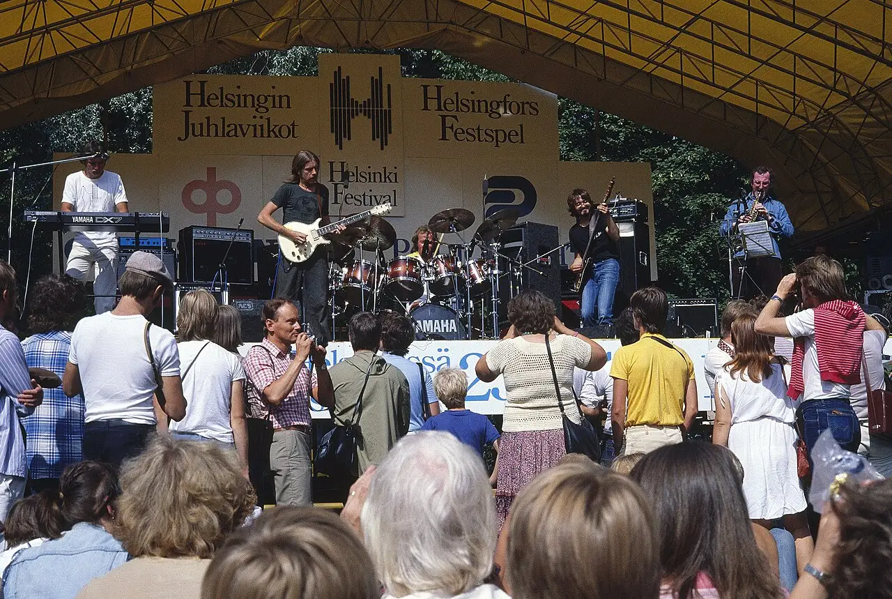 Banda tocando num palco ao ar livre sob a faixa do Helsingin Juhlaviikot, com o público de costas em primeiro plano.