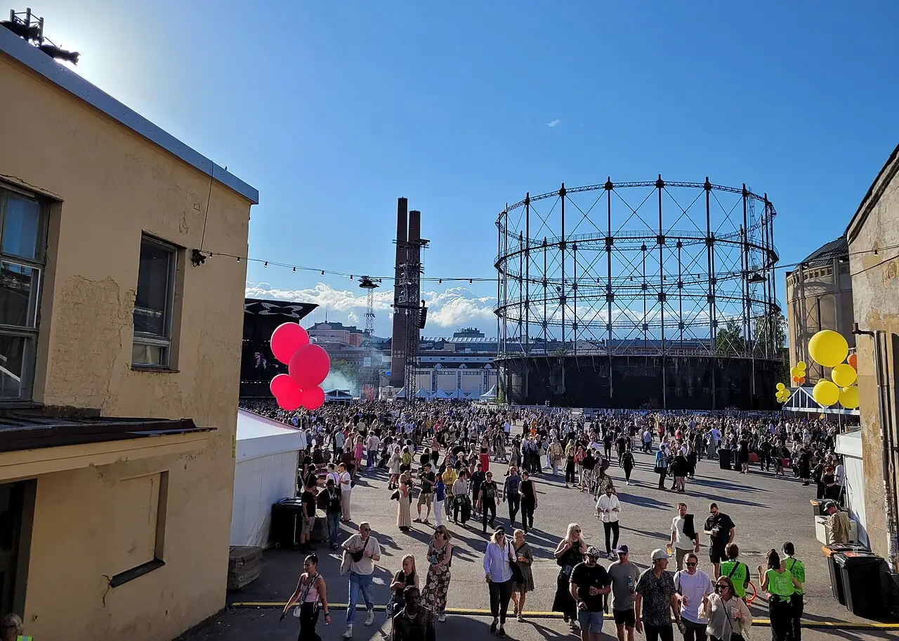 Multidão caminhando pelo recinto do Flow Festival em Suvilahti, sob céu azul, com o gasômetro de estrutura metálica ao fundo, em agosto de 2023.