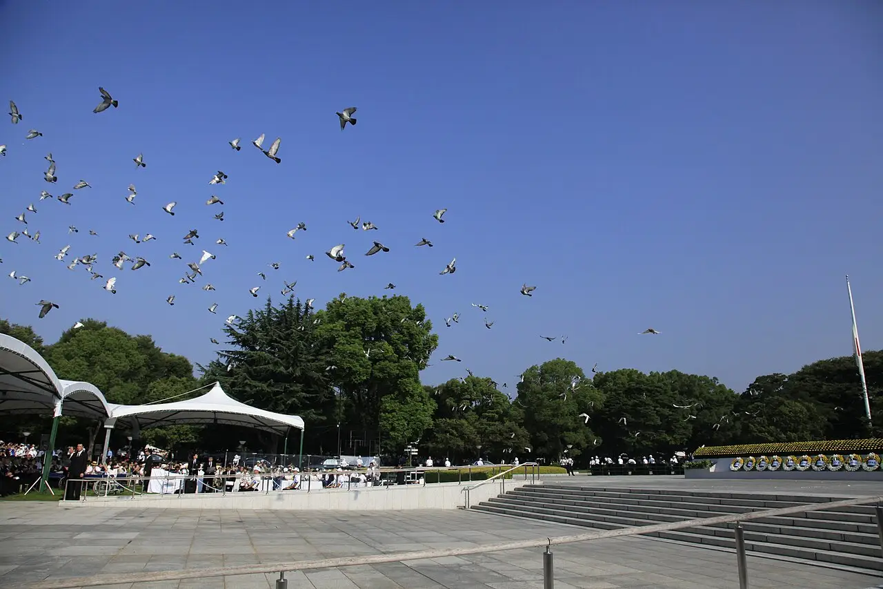 Pombas voando sobre o Parque Memorial da Paz de Hiroshima, com tendas brancas e árvores ao redor da área da cerimônia.