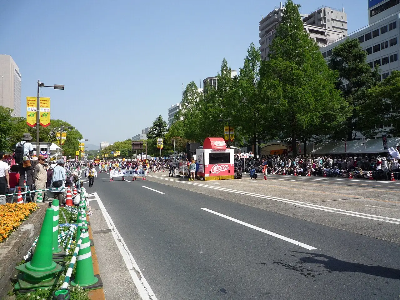 Avenida larga de Hiroshima bordada por árvores e canteiros de flores, com espectadores na calçada e um carro alegórico ao longe, em 2011.