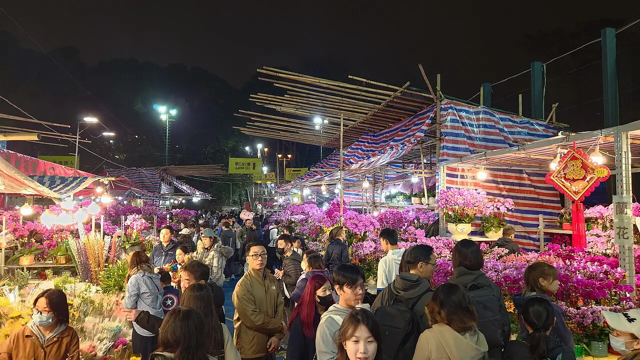 Multidão entre bancas de orquídeas rosa e roxas sob toldos listrados, à noite, na feira do Ano-Novo Lunar em Hong Kong.