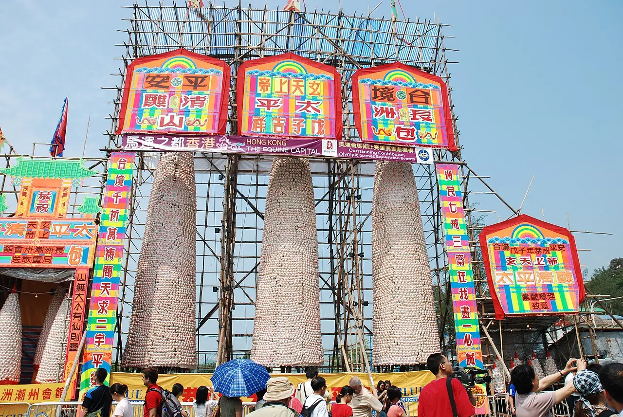 Torres de bambu cobertas de pães, com painéis coloridos no alto e público ao redor, no Festival dos Pães de Cheung Chau.