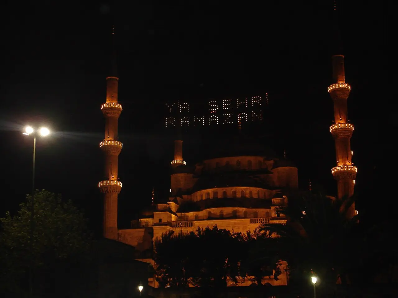 Mesquita Azul iluminada à noite, com uma mensagem luminosa suspensa entre dois minaretes durante o Ramadã.