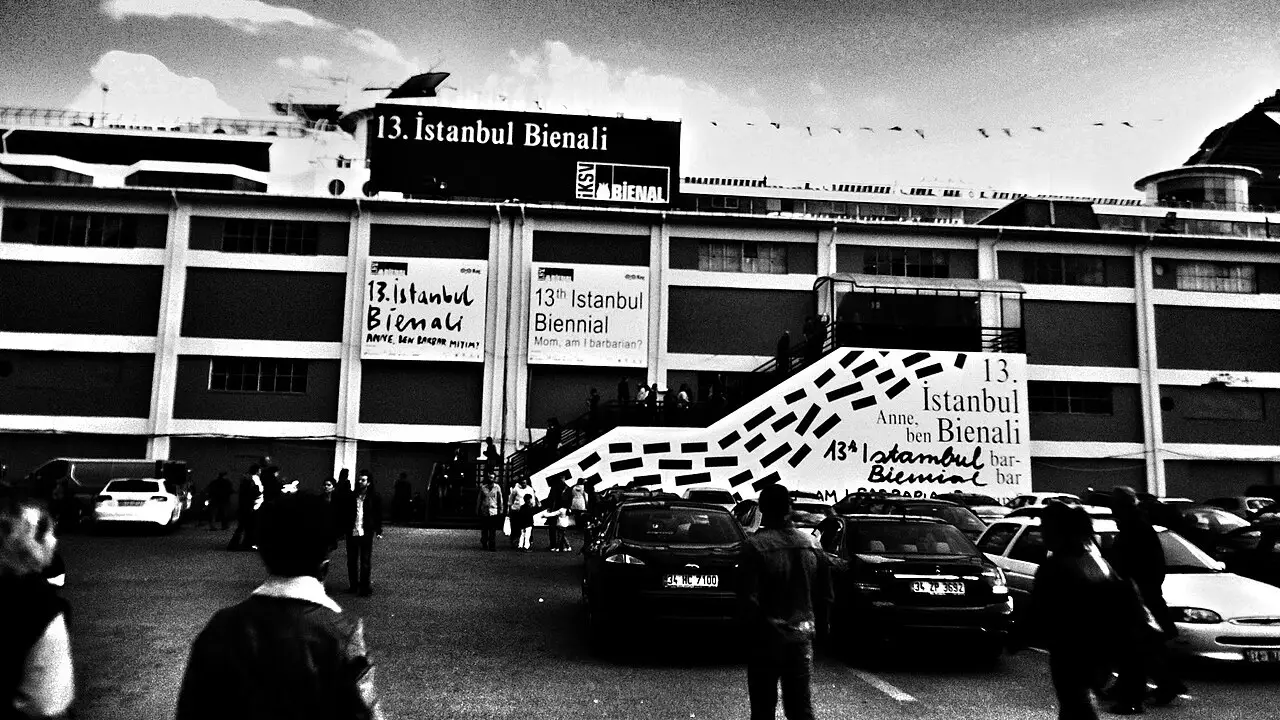 Prédio de vários andares com faixas da 13ª Bienal de Istambul na fachada e carros estacionados na frente, em foto em preto e branco.