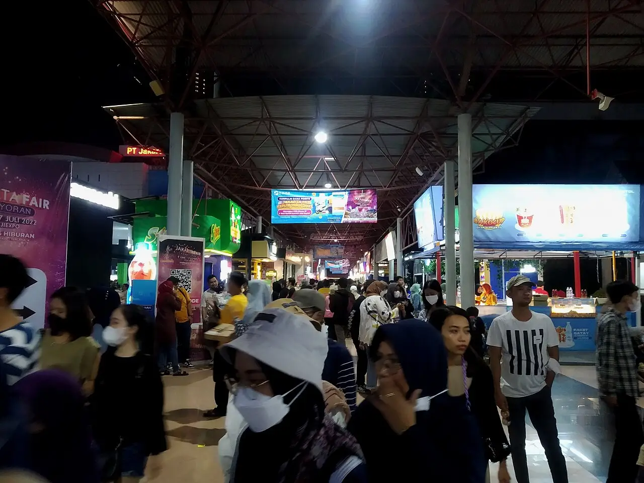 Multidão de visitantes, alguns de máscara, circulando à noite sob uma cobertura entre estandes iluminados na Jakarta Fair de 2022.