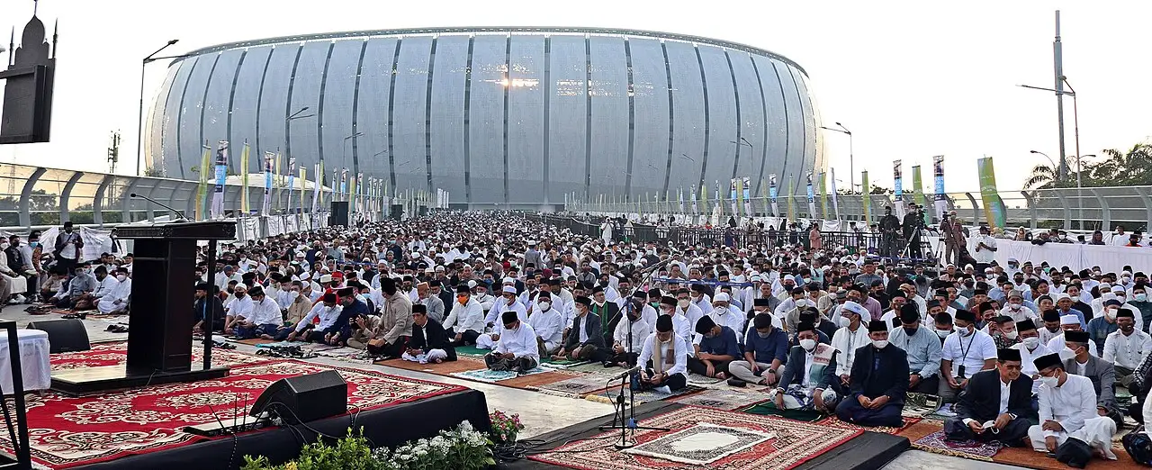 Fiéis de branco sentados em fileiras diante de um púlpito, em oração do Idul Fitri ao ar livre, com o Estádio Internacional de Jacarta ao fundo.
