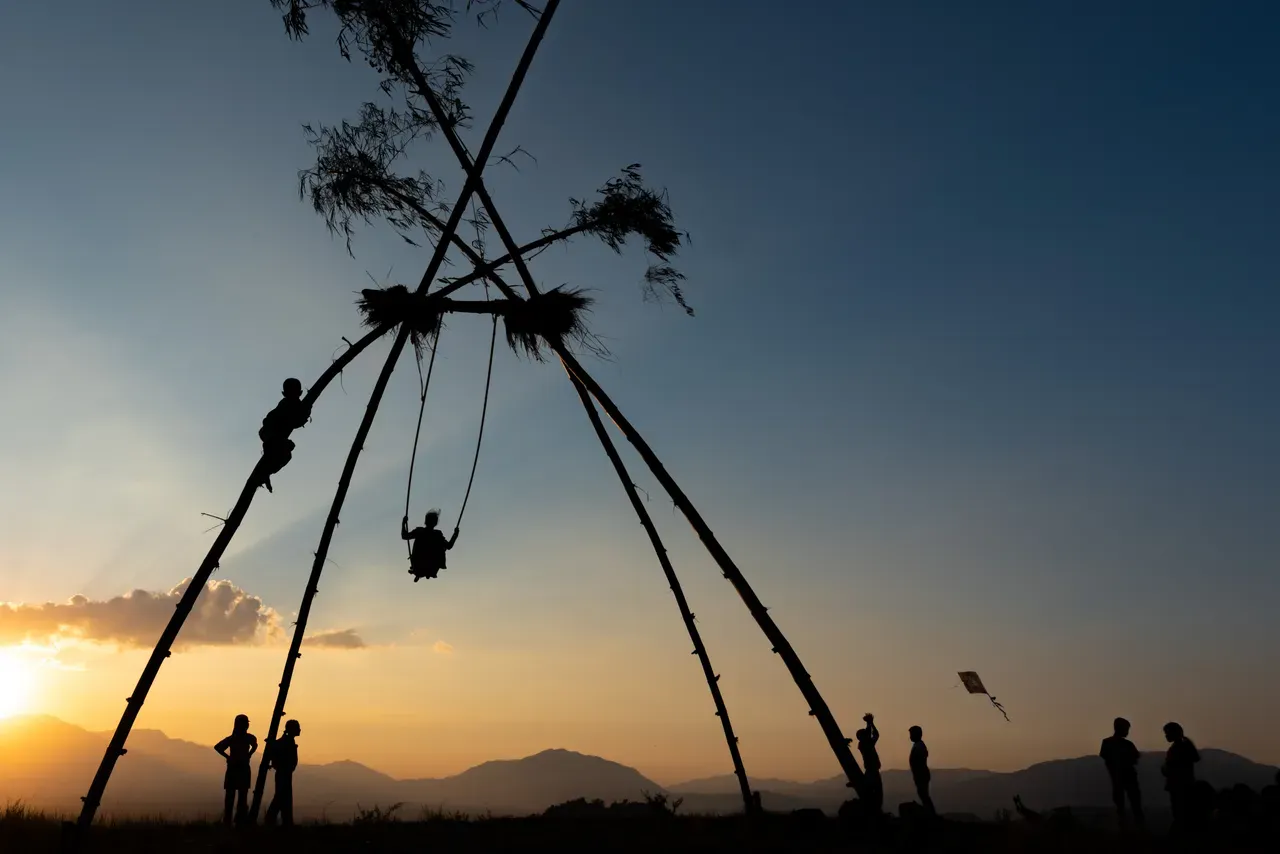 Silhuetas em volta de um balanço alto de bambu, ao pôr do sol, com uma pipa no céu e morros atrás, no Dashain de 25 de outubro de 2023, nos arredores do vale de Katmandu.