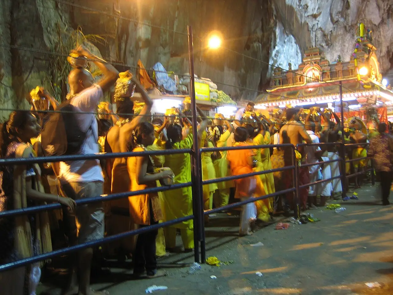 Devotos em roupas amarelas e laranja enfileirados atrás de grades sob luzes, dentro das Cavernas Batu, durante o Thaipusam.