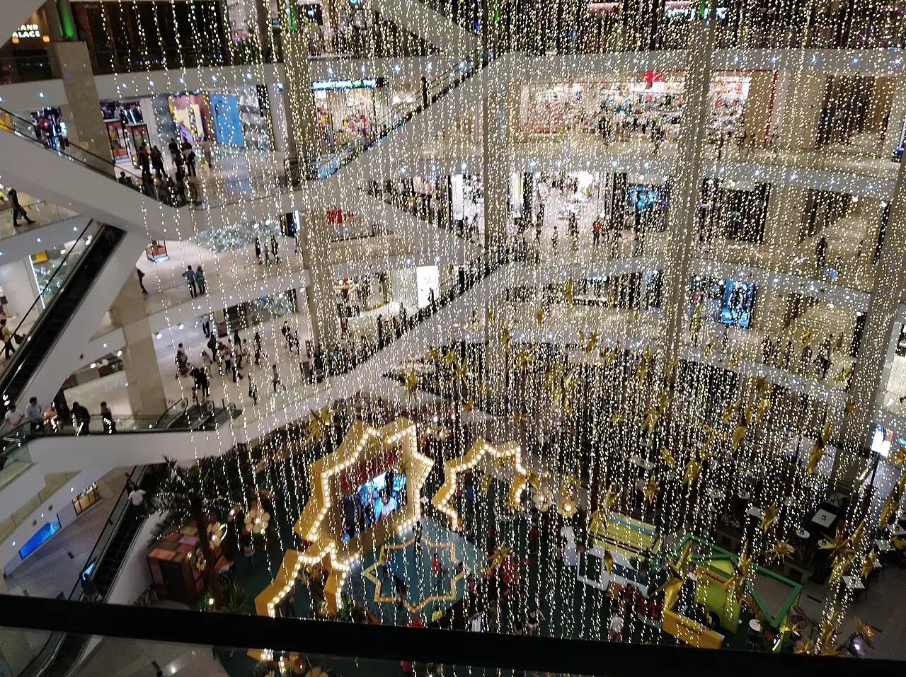 Átrio de shopping em vários andares decorado com cortinas de luzes e formas iluminadas para o Hari Raya Aidilfitri, visto de cima.