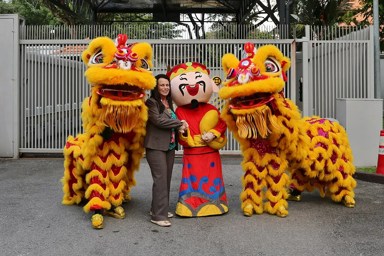 Dois leões dourados e vermelhos e um mascote de traje vermelho posando com uma pessoa diante de um portão, em festa de Ano-Novo Chinês em 2018.