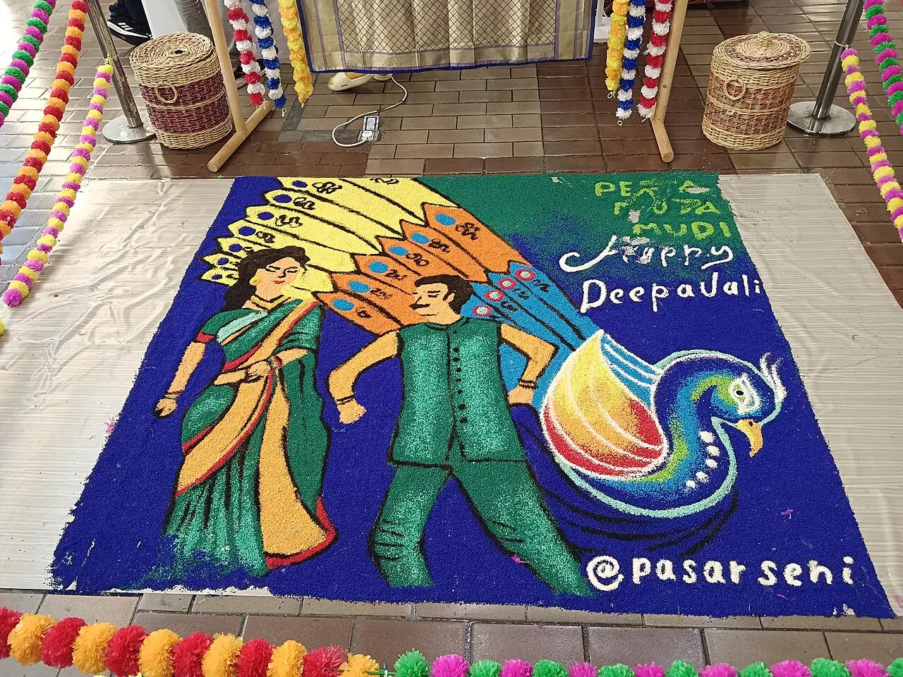 Grande kolam de arroz colorido com duas figuras e um pavão, e a inscrição Happy Deepavali, exposto no Pasar Seni.