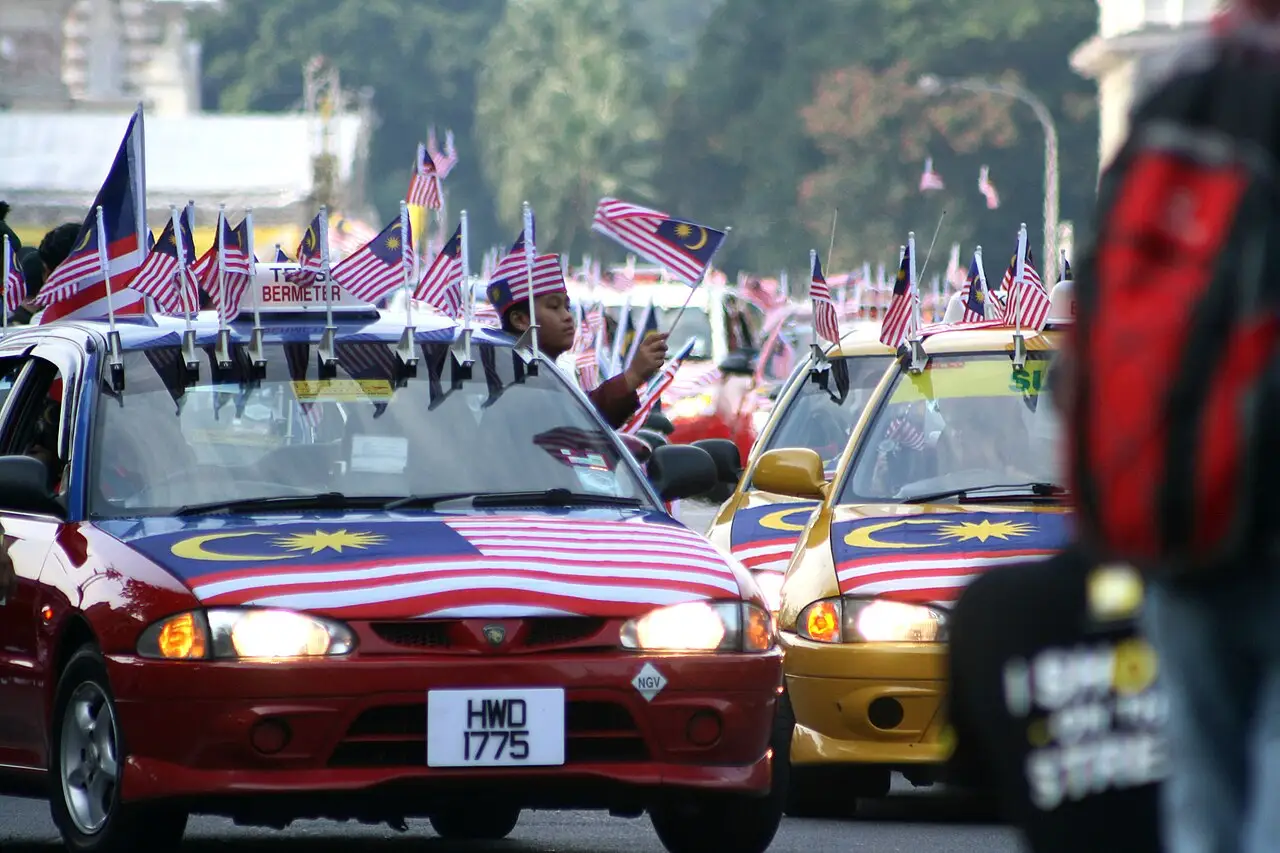 Táxis e carros cobertos por bandeiras da Malásia desfilando em meio à multidão em Dataran Merdeka, nas celebrações de 2011.
