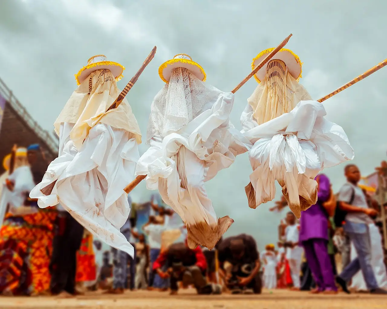 Três mascarados Eyo vestidos de branco, com chapéus e varas, dançando diante de espectadores, no festival Eyo de Lagos em 2025.