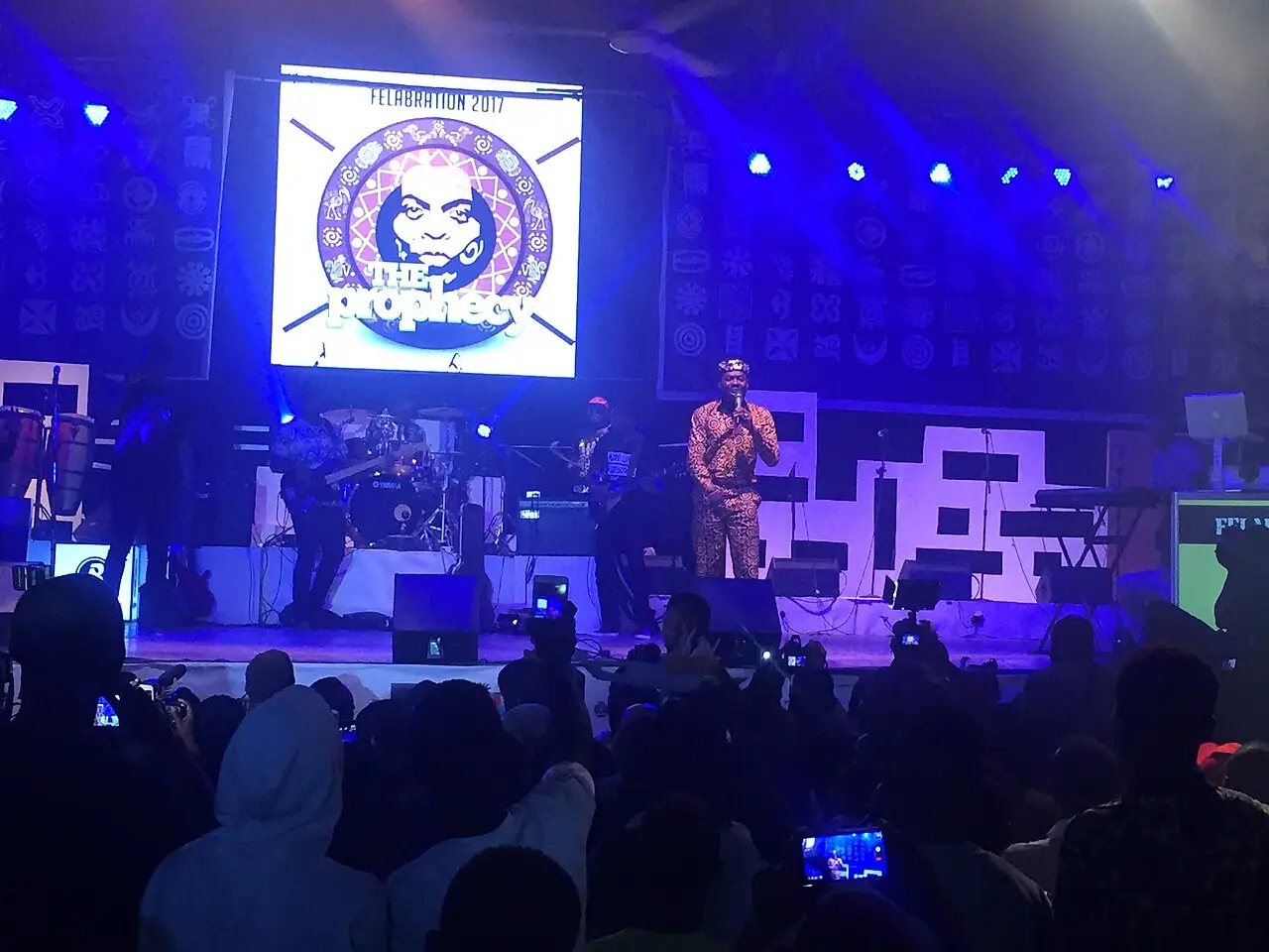 Artista em pé num palco iluminado em azul diante de um telão com o emblema do Felabration, com a plateia à sombra em primeiro plano.
