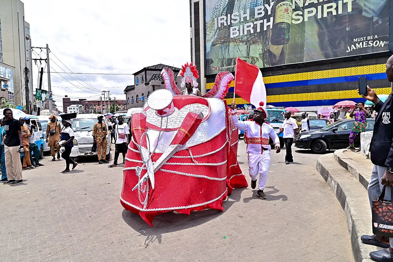 Figura de traje vermelho e branco, largo e amarrado com cordas, andando por uma rua de Lagos entre espectadores durante o Carnaval Fanti de 2026.