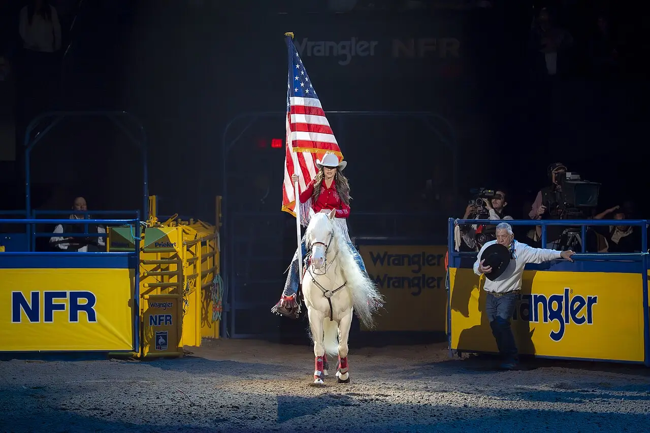 Amazona num cavalo branco carregando a bandeira dos Estados Unidos na arena do National Finals Rodeo, entre barreiras com o logotipo NFR.
