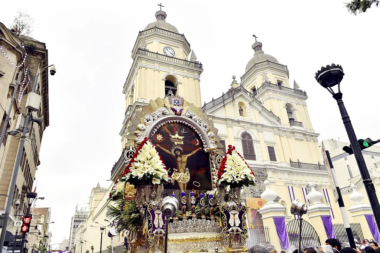 Andor com a imagem do Señor de los Milagros, coberto de flores brancas, diante de uma igreja de torres brancas em Lima.