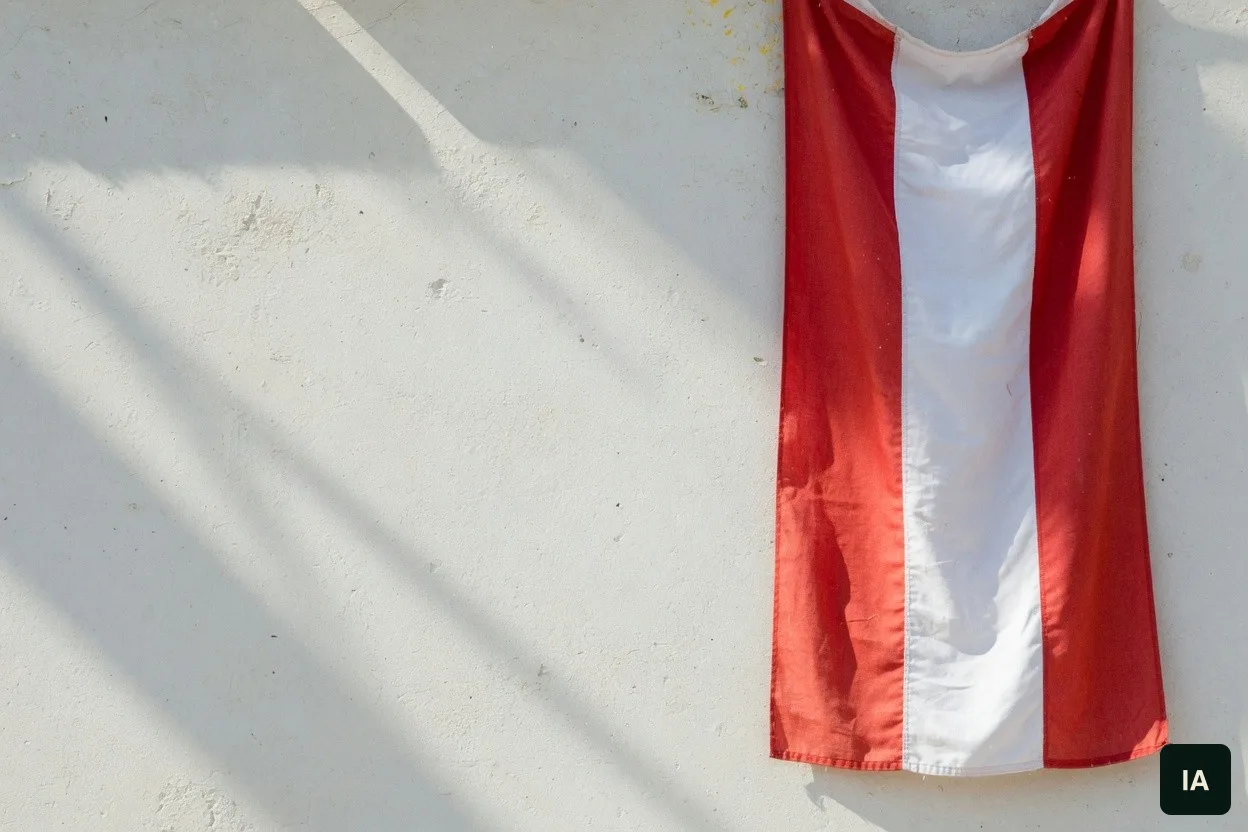 Bandeira em três faixas verticais, vermelho, branco e vermelho, sobre uma parede clara, ao sol.