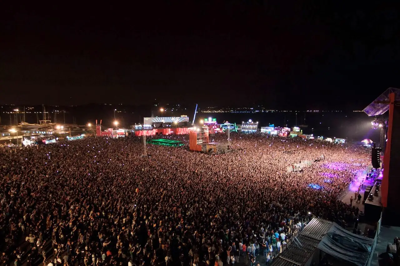 Vista de cima de uma multidão enorme diante do palco à noite, com luzes coloridas e o rio ao fundo, no festival de música de Algés.