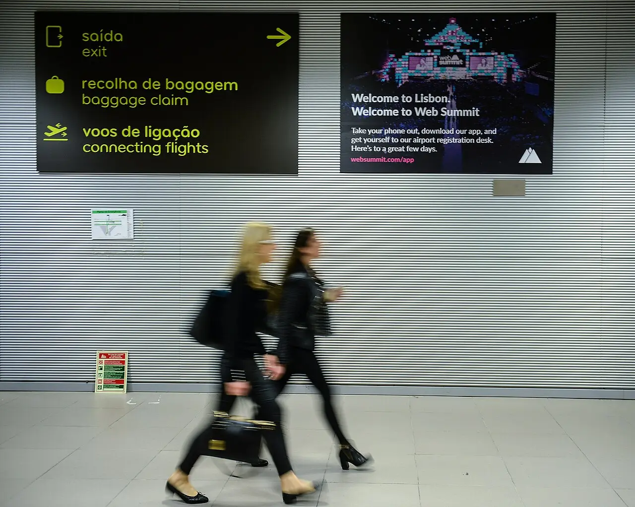 Painel de boas-vindas ao Web Summit no aeroporto de Lisboa, acima de duas passageiras em movimento, em novembro de 2017.