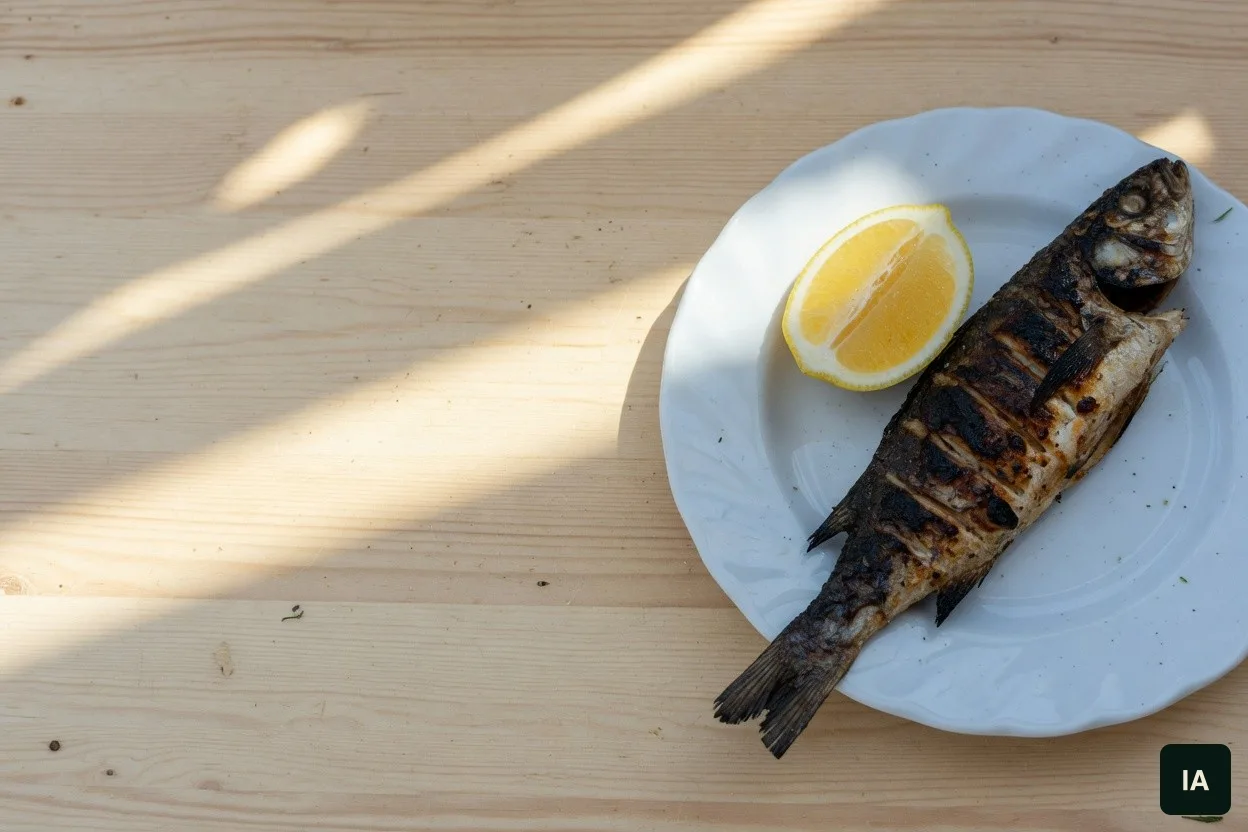 Peixe grelhado inteiro e um gomo de limão num prato branco, numa mesa clara ao sol.