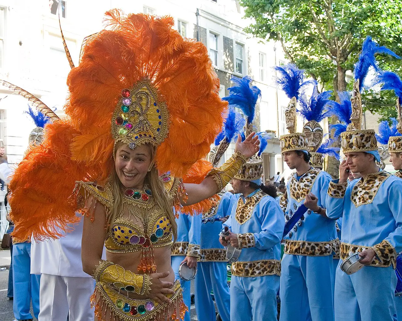 Dançarina com grande cocar de plumas laranja e dourado desfilando ao lado de integrantes de azul, no Notting Hill Carnival de 2006.