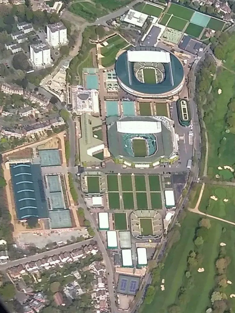 Vista aérea do complexo de Wimbledon, com duas grandes quadras cobertas e várias quadras verdes menores entre caminhos e edifícios.