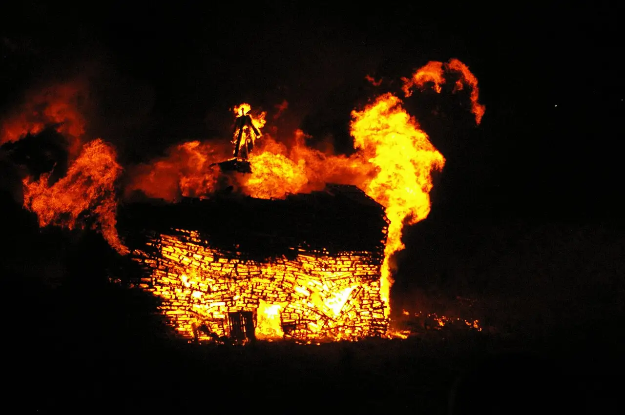 Grande fogueira acesa à noite no Carshalton Park, com chamas altas e pessoas ao fundo, na Bonfire Night.