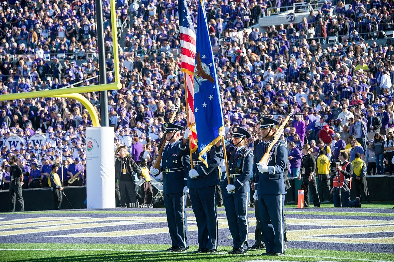 Três militares de uniforme azul, com a bandeira dos Estados Unidos e uma bandeira azul, no gramado do Rose Bowl, diante da arquibancada, em 1º de janeiro de 2019.