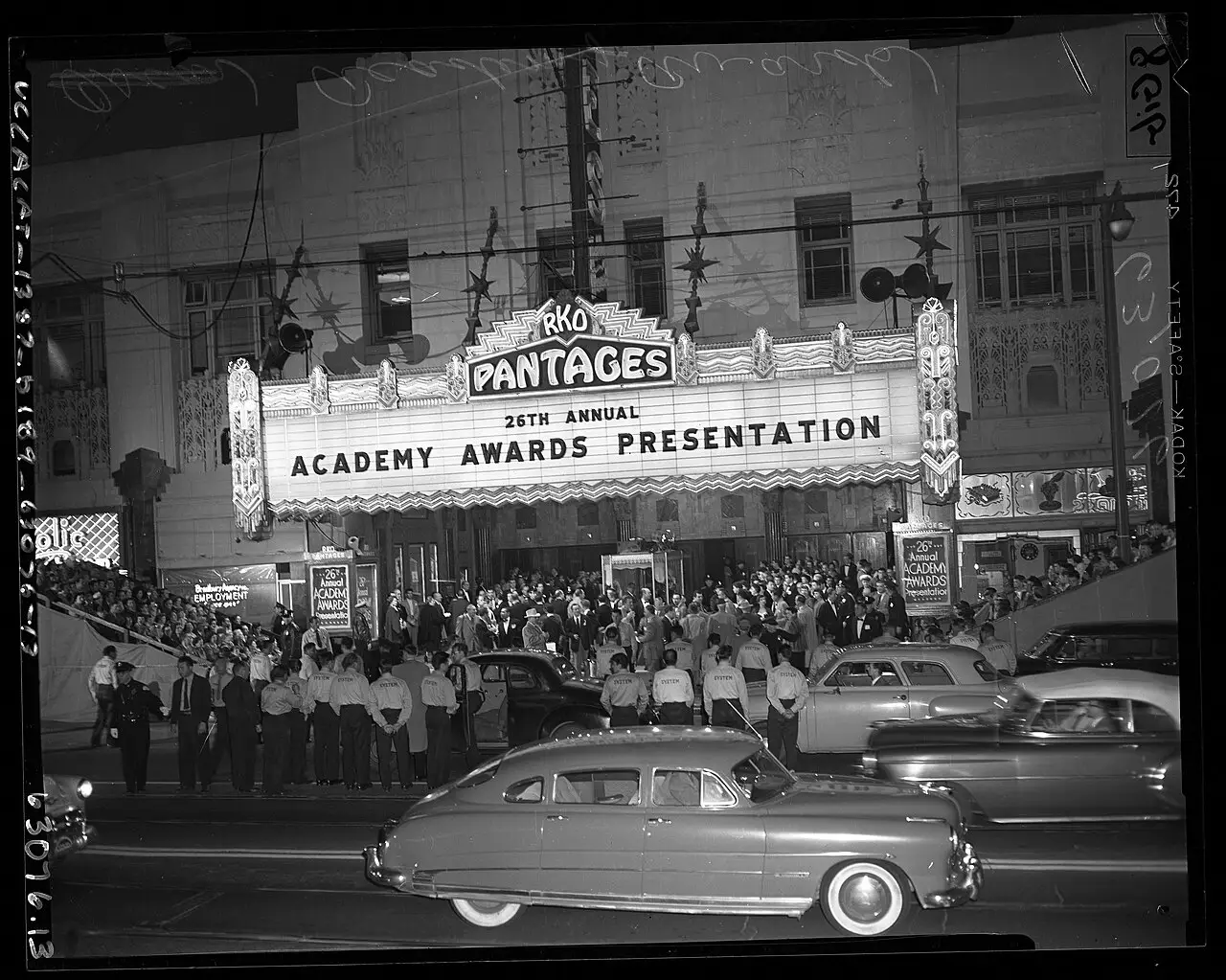 Fachada do RKO Pantages Theater com o letreiro da 26ª entrega do Oscar, multidão diante e carros estacionados na rua, em 1954.