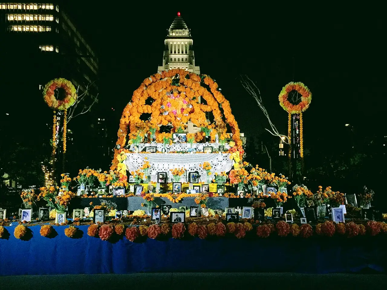 Grande altar decorado com flores laranja e velas, à noite, no Grand Park de Los Angeles, com a prefeitura iluminada ao fundo.