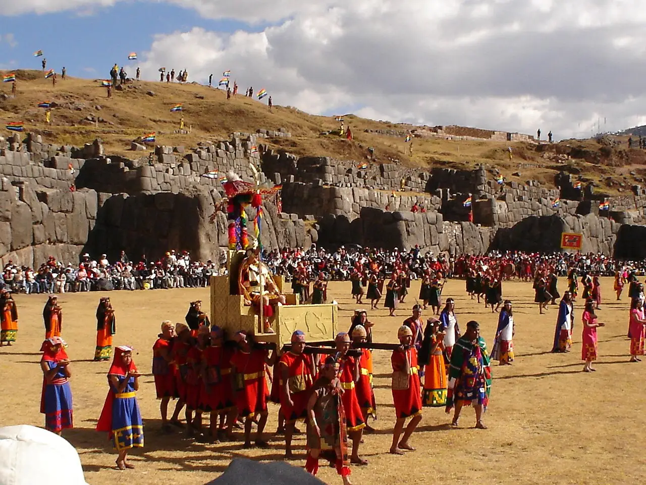 Figura do Inca carregada num andor por participantes de trajes vermelhos diante dos muros de pedra de Sacsayhuamán, com público na encosta.