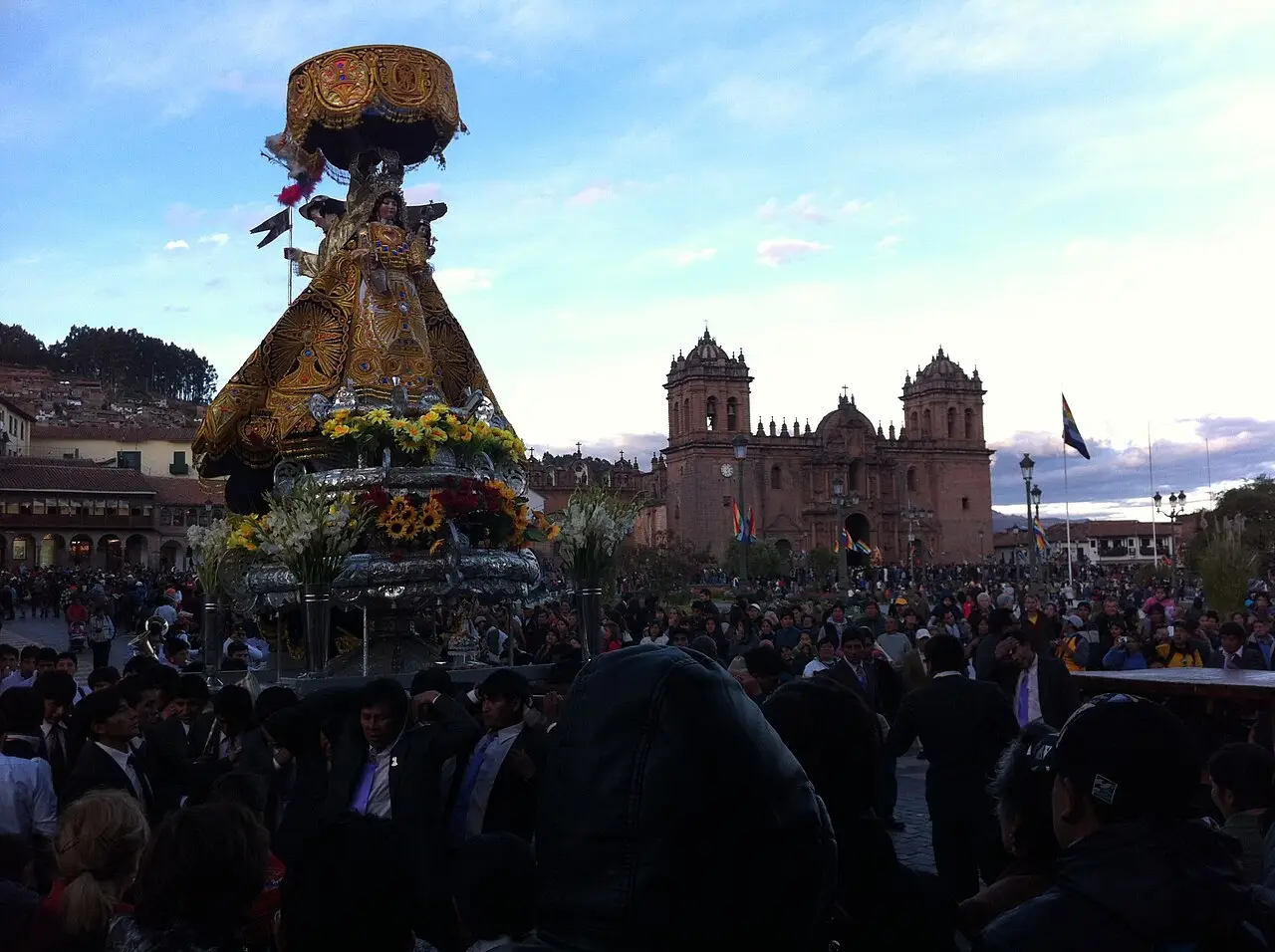 Grande imagem em andor com dossel dourado, cercada por multidão, com a catedral de Cusco ao fundo, na festa da Plaza de Armas.