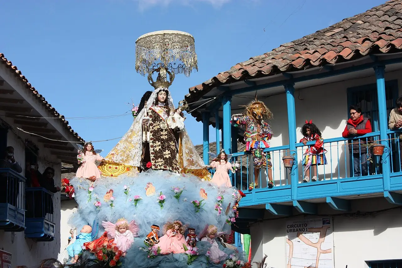Imagem da Virgen del Carmen sobre um andor florido e com dossel, diante de uma casa de balcão azul, em Paucartambo.