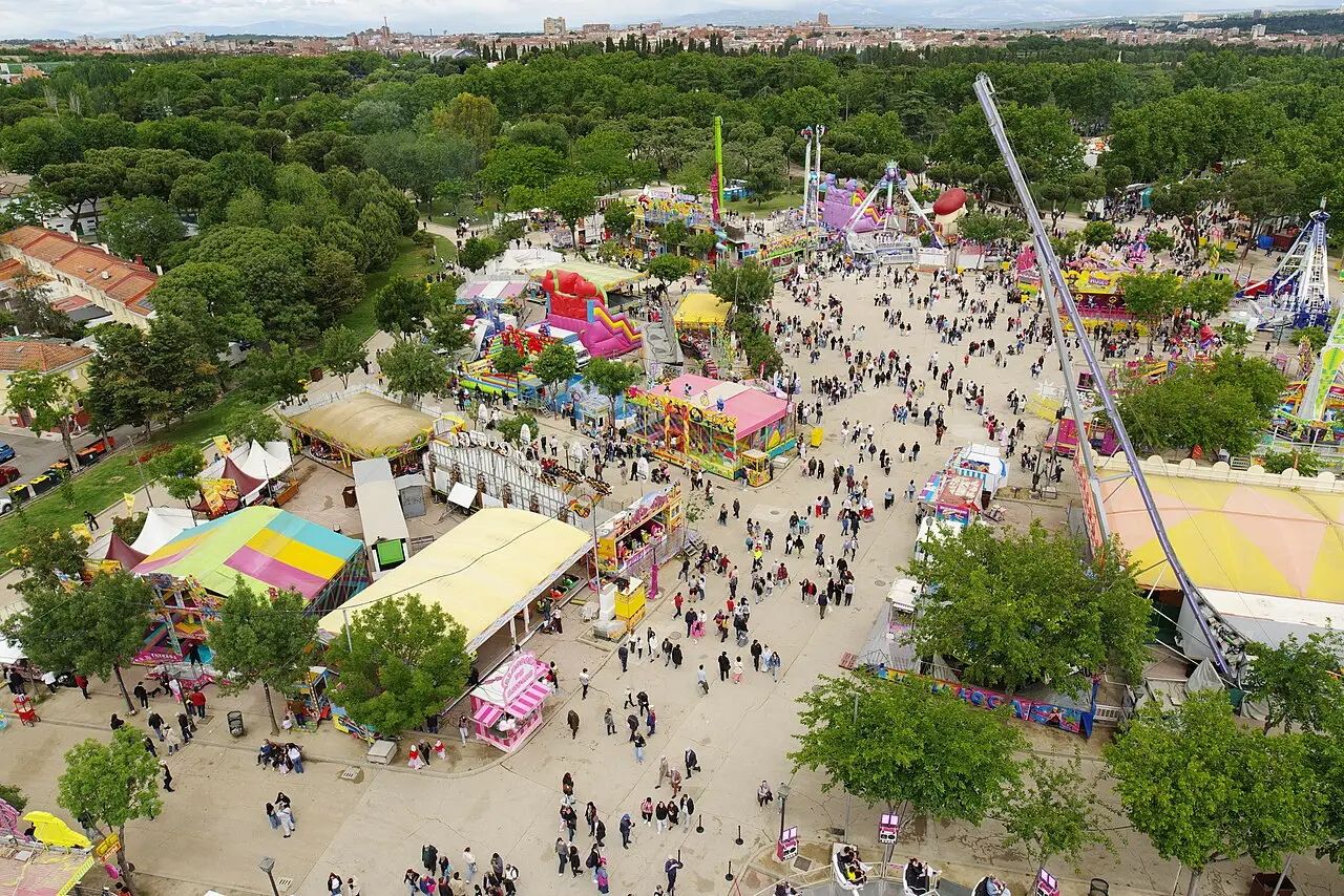 Vista de cima do parque de diversões da Fiesta de San Isidro em Madri, em 2026, com barracas coloridas e visitantes entre as árvores.