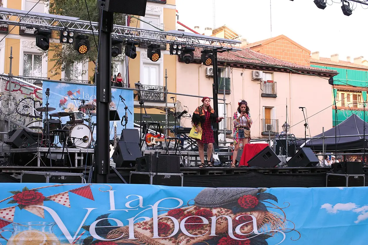 Duas pessoas cantam num palco de rua, com bateria atrás e uma faixa azul onde se lê la Verbena.