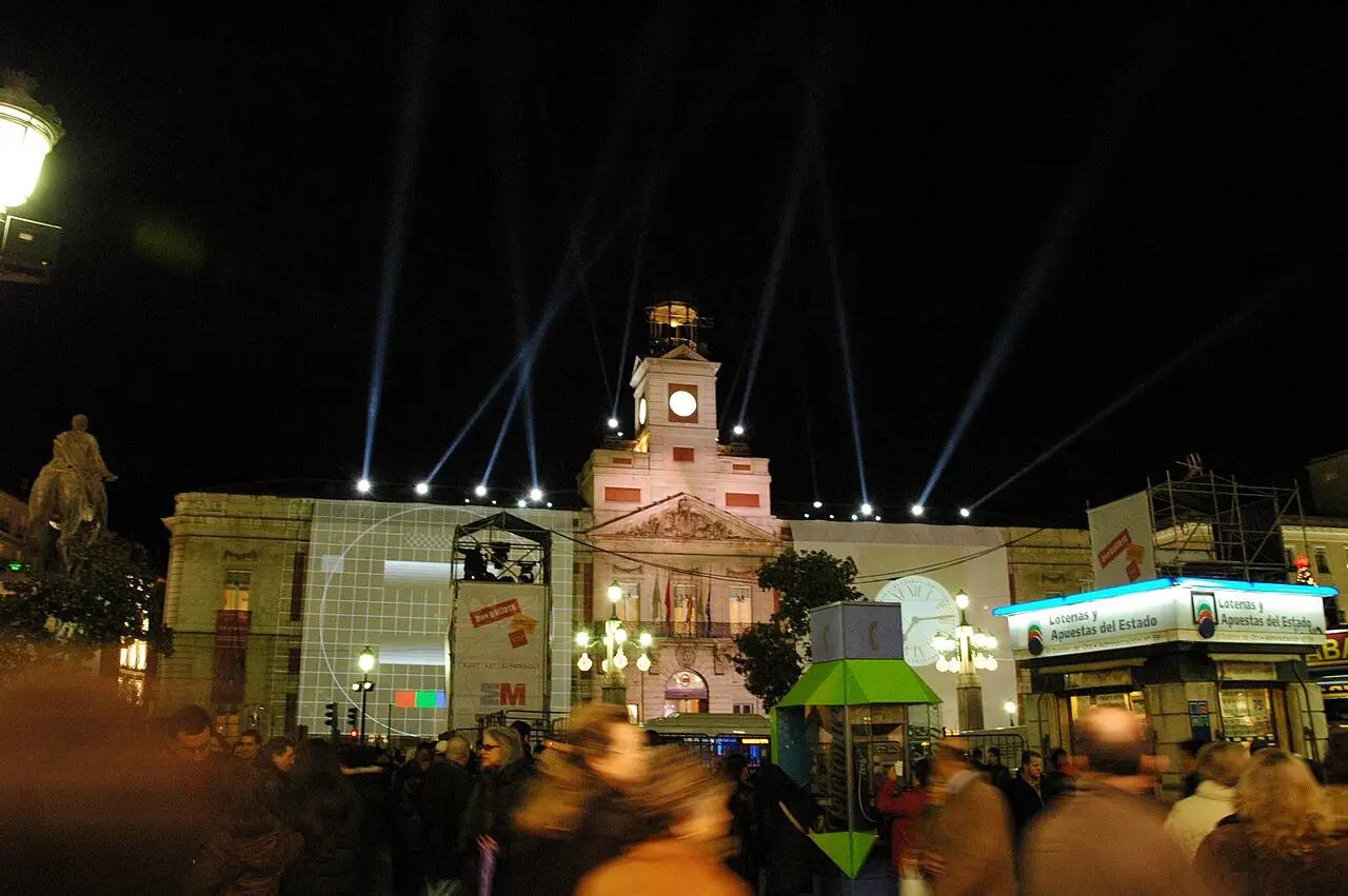 Feixes de luz saem do prédio do relógio, e a praça à frente está cheia de gente.