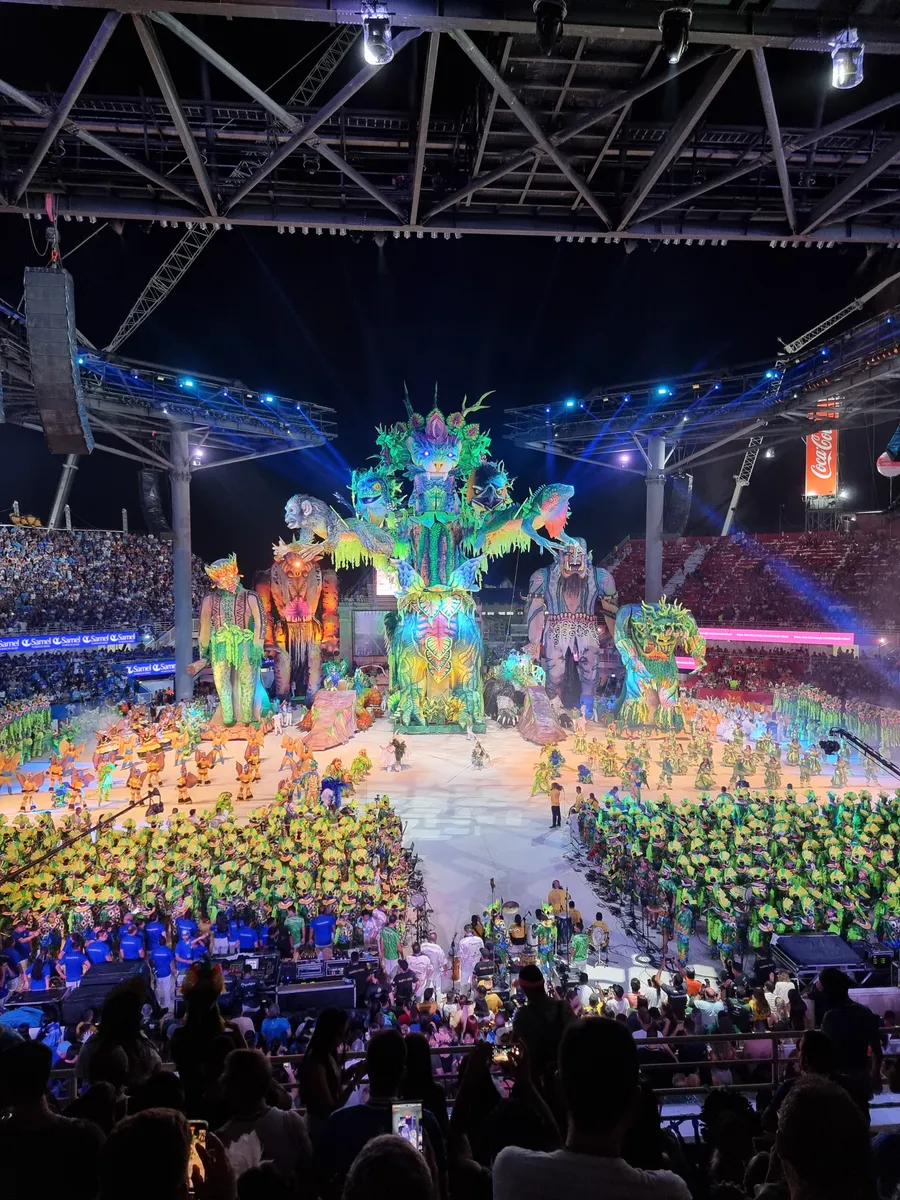 Alegorías iluminadas en el centro del Bumbódromo, con bloques de participantes de verde y de azul y el público visto de espaldas.