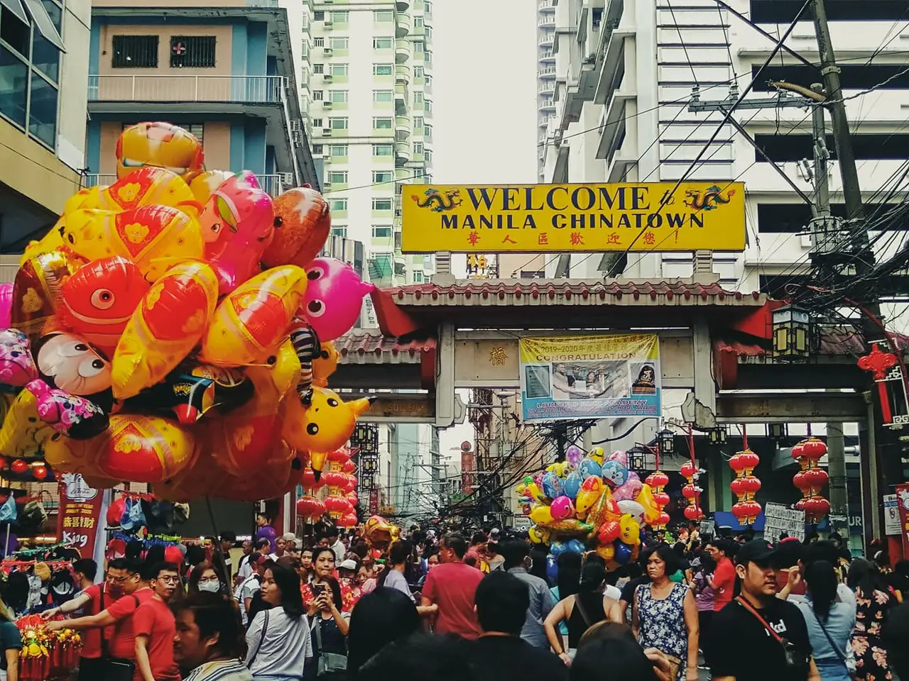 A rua está cheia sob o letreiro Welcome to Manila Chinatown, com balões de peixe laranja e lanternas vermelhas.