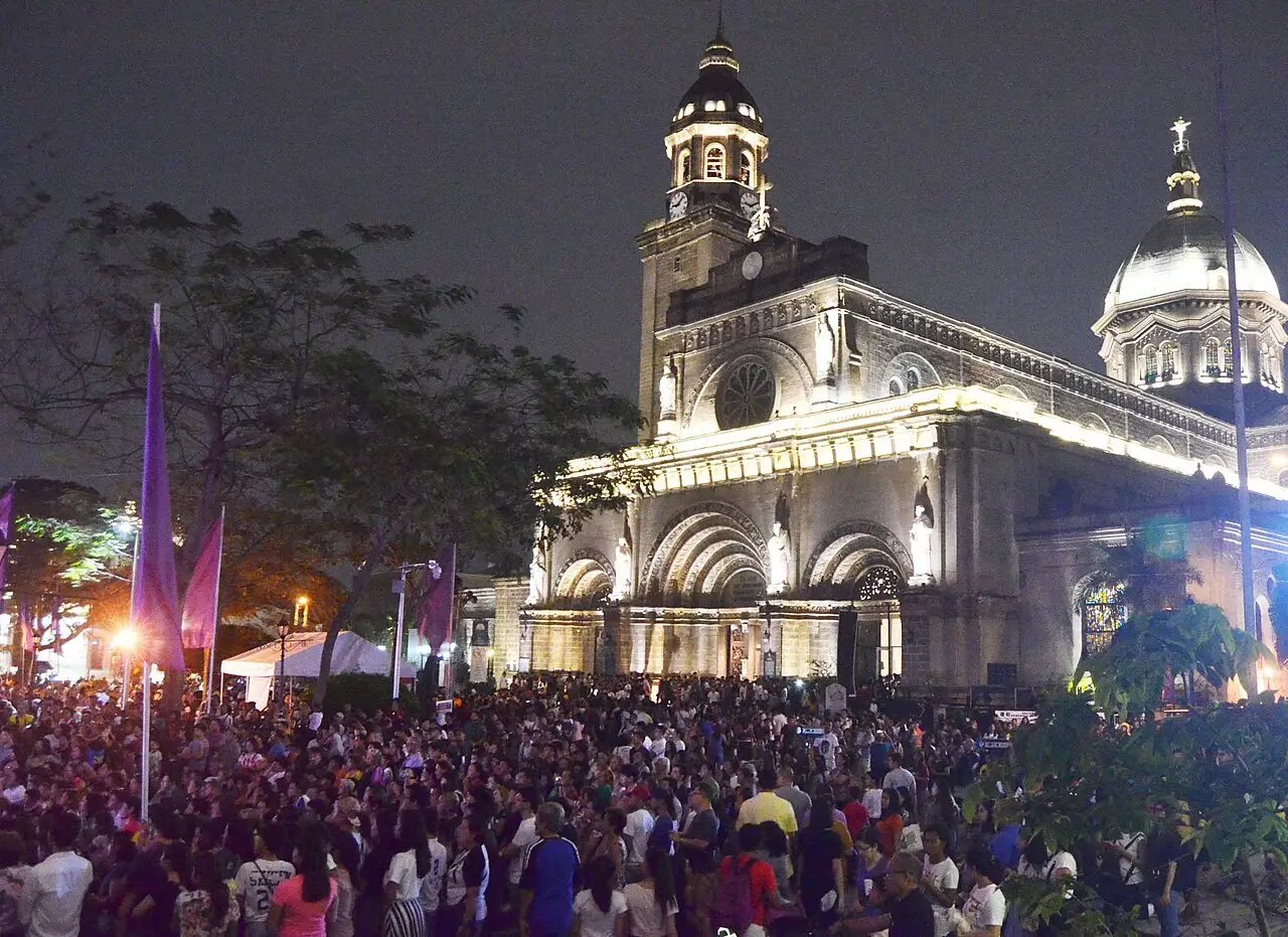 Uma multidão ocupa a praça diante da catedral iluminada, de noite, com bandeiras roxas à esquerda.