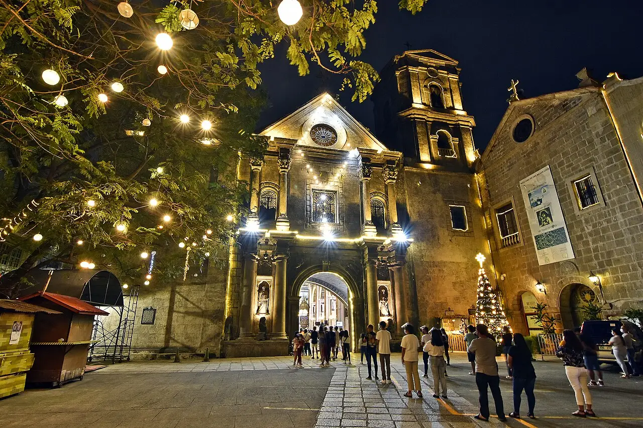 Pessoas estão na porta da igreja de pedra, à noite, sob fios de lâmpadas redondas.
