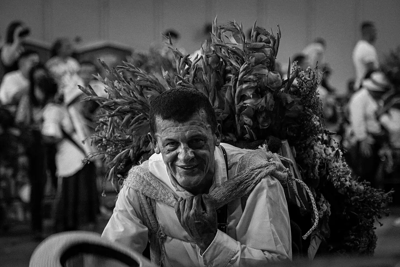 Em preto e branco, um homem sorri com um arranjo grande de flores e folhas nas costas, e há gente atrás.