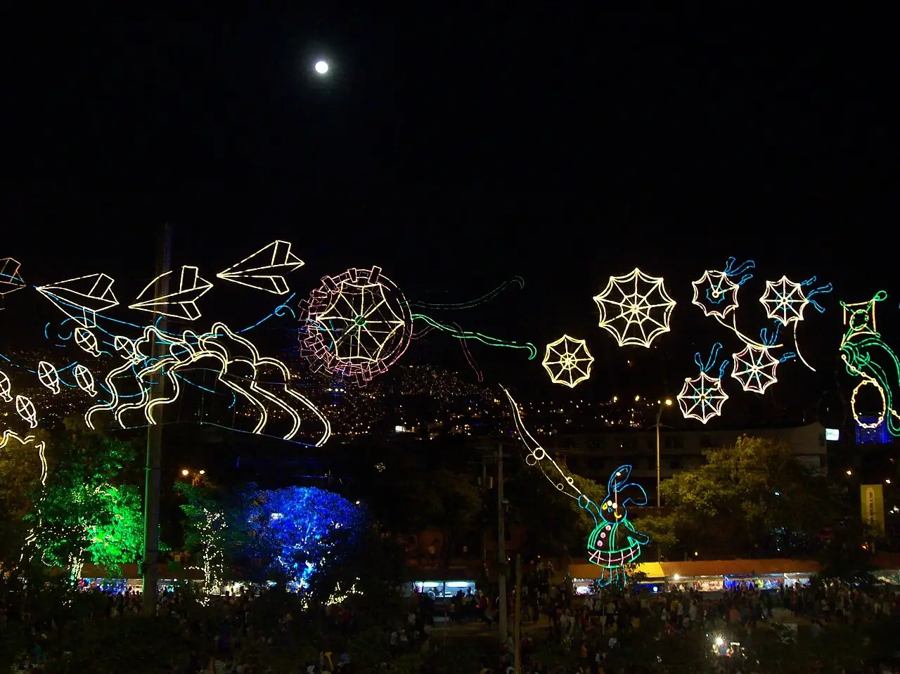 Figuras de luz em forma de flocos e flores atravessam o parque à noite, com árvores verdes e azuis e gente embaixo.
