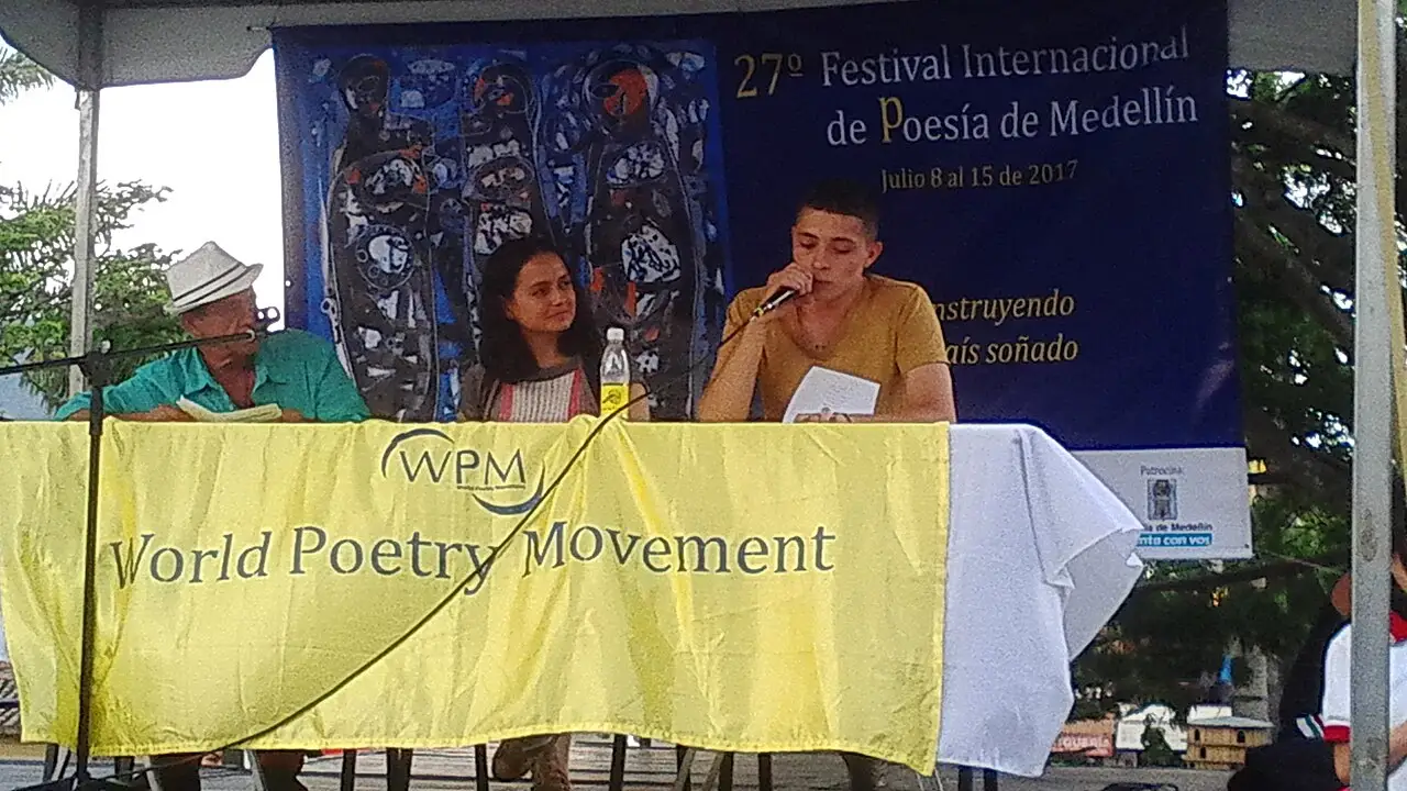 Três pessoas estão sentadas a uma mesa sob o cartaz do 27º Festival Internacional de Poesía de Medellín, com uma faixa amarela à frente.