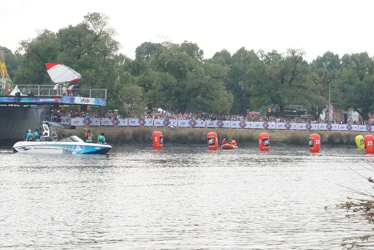 Barcos e boias vermelhas e brancas estão no rio, com gente na margem e uma plataforma escura ao fundo.